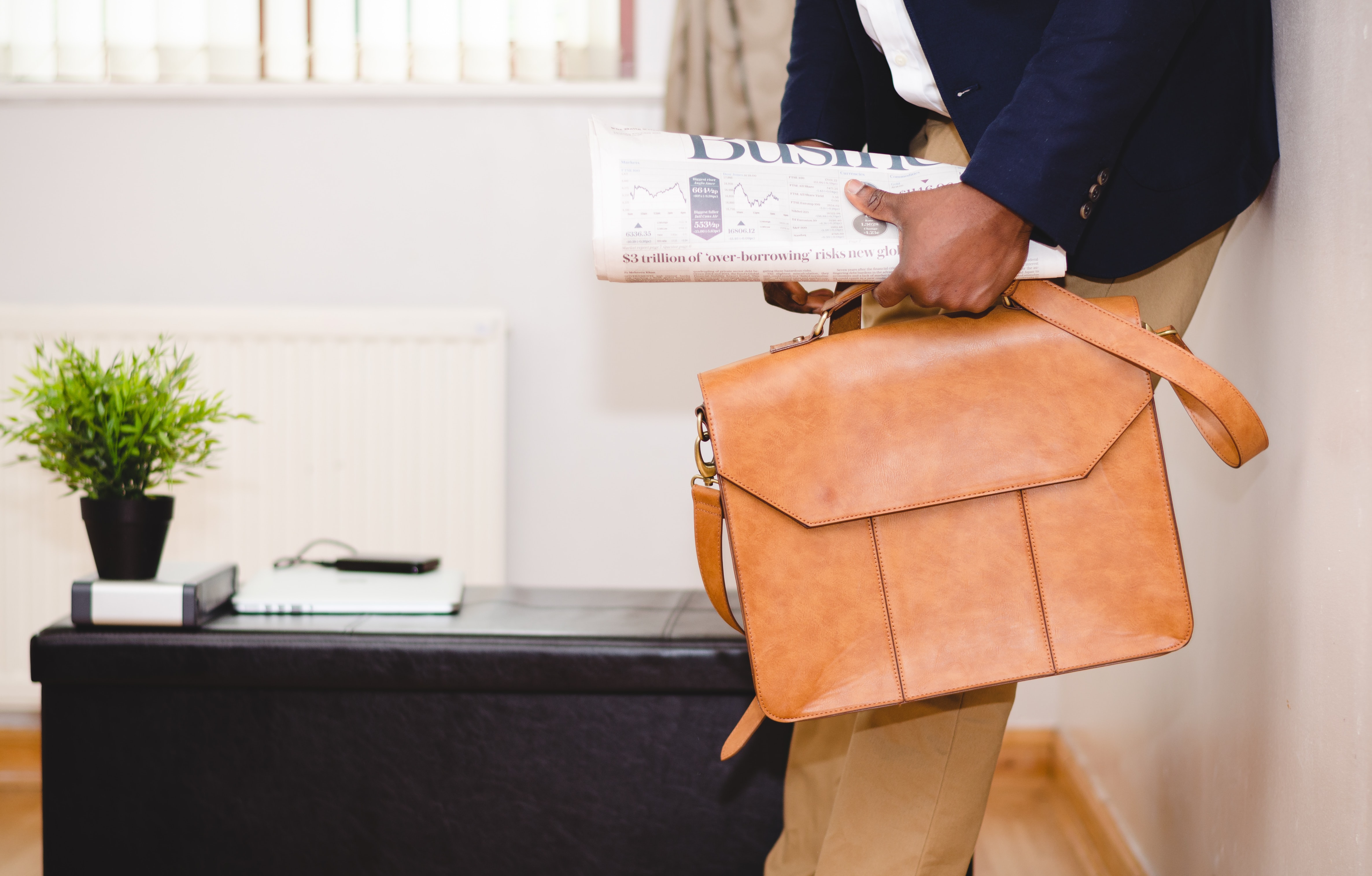 a man with dark skin shown in profile shin to shoulder carries a light brown leather briefcase past an ottoman with a potted plant and office objects