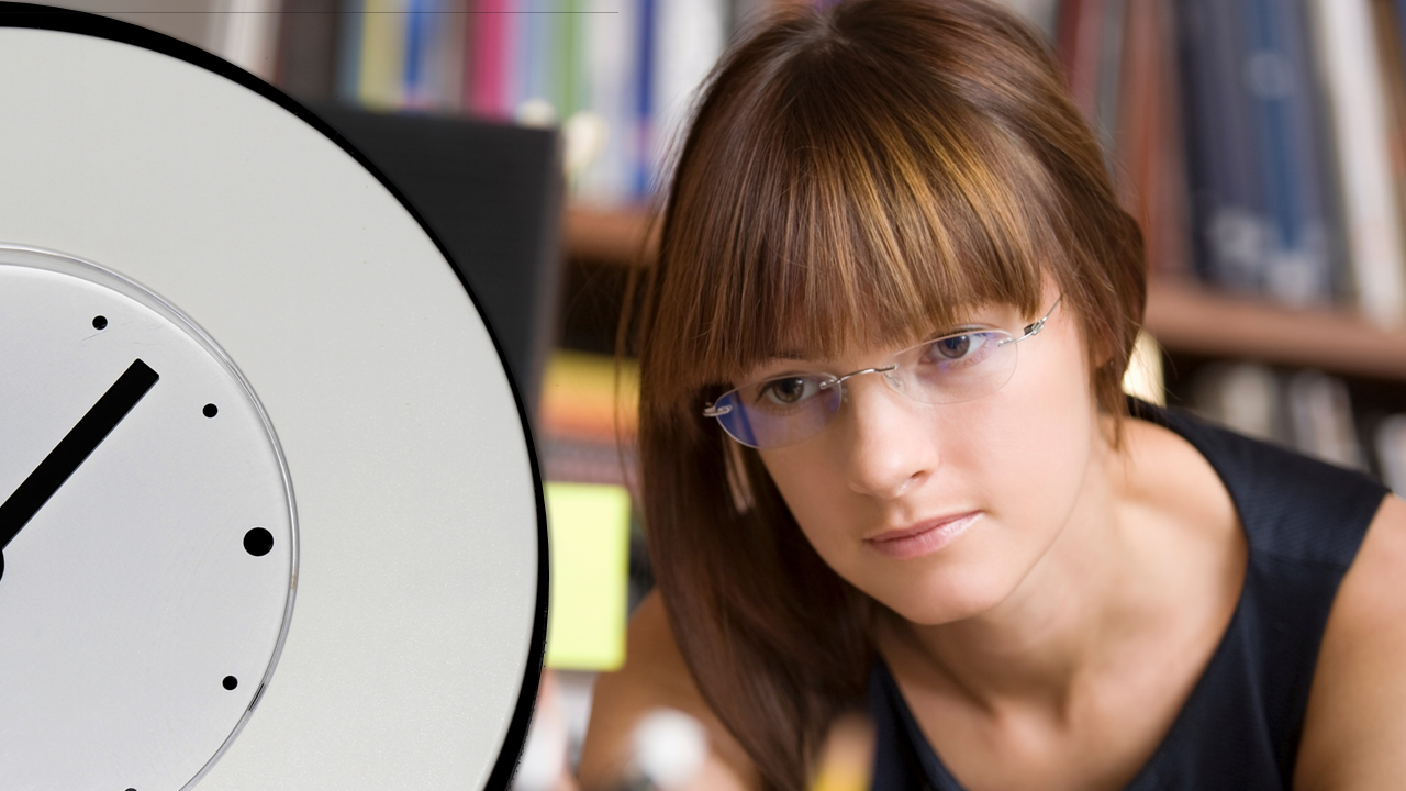 A person with bangs and glasses looks at a computer with a clock in front of it.