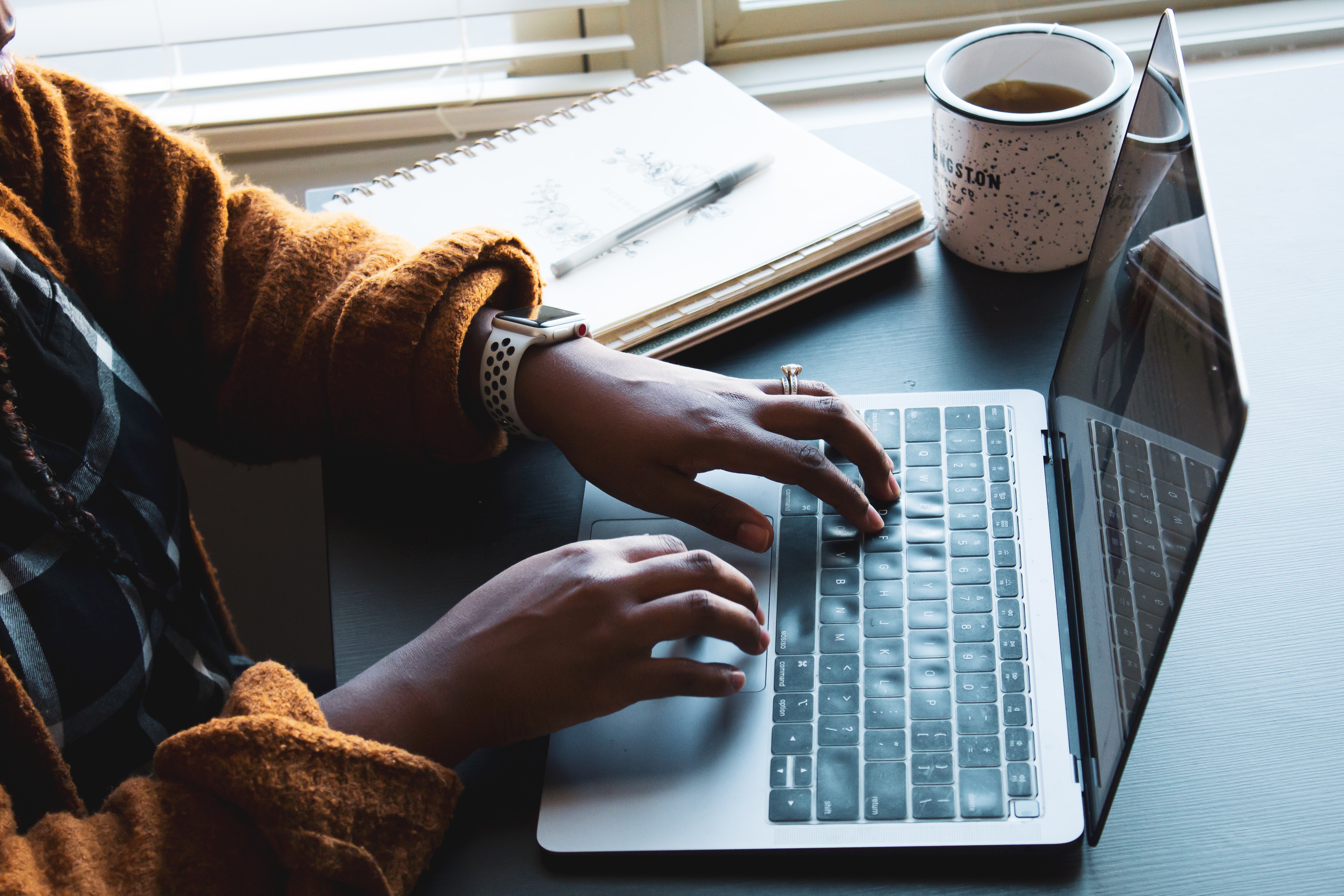 View of a desk where someone is typing on a laptop by a window with a notebook and coffee mug sitting nearby