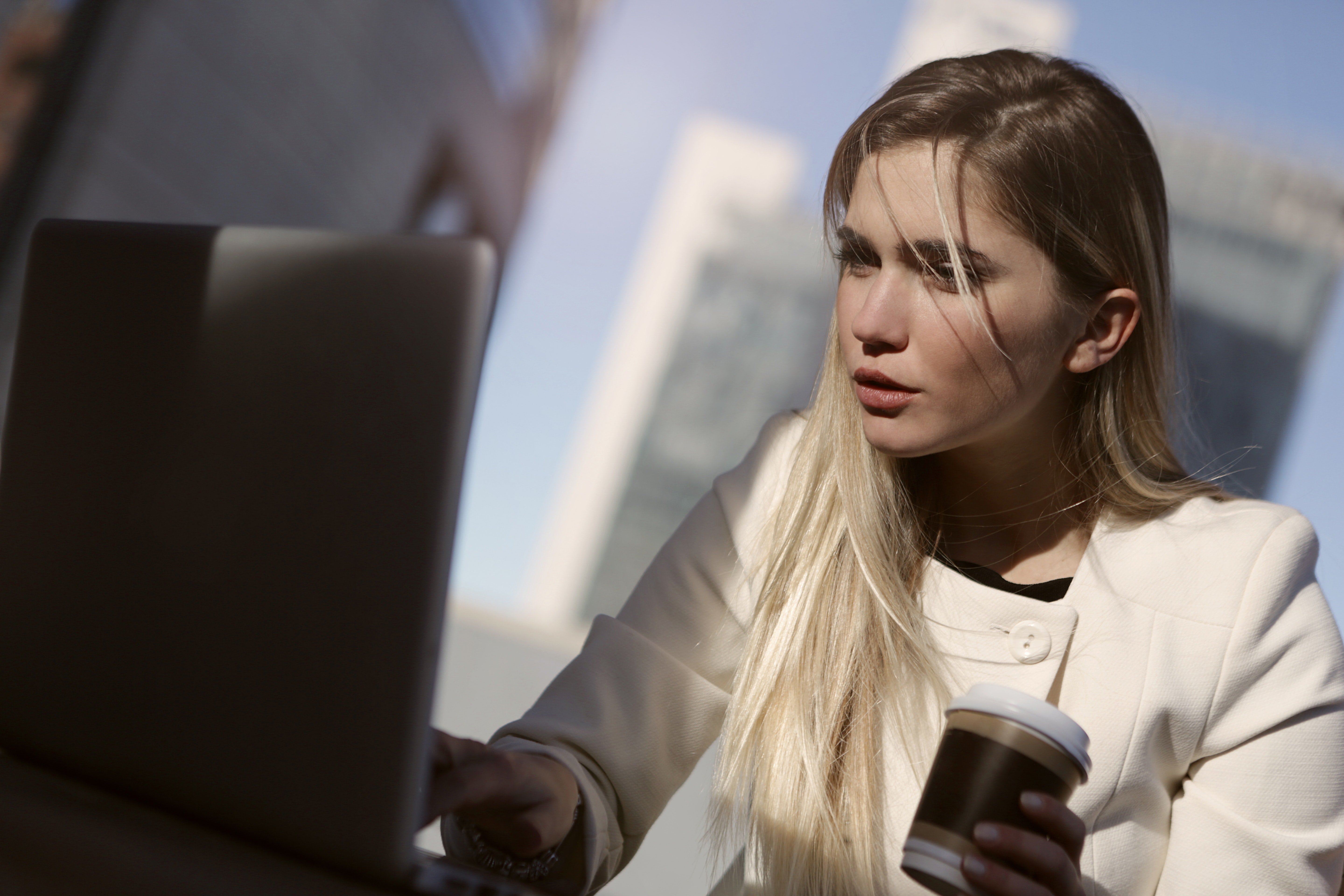 A light skinned blonde woman with long hair sits as a computer looking concerned while holding a disposable coffee cup