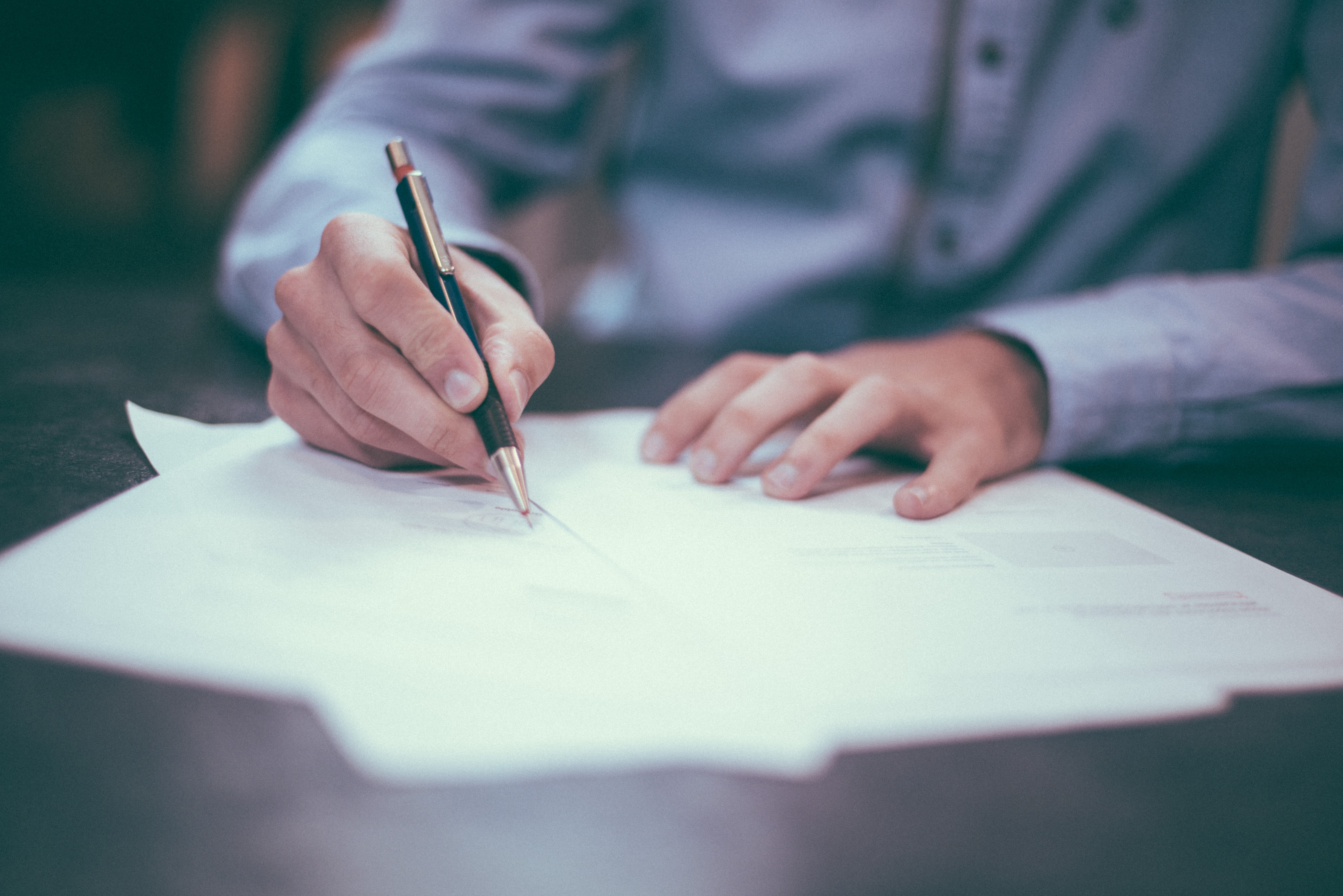 man with light skin wearing button down shirt writes on a pile of documents