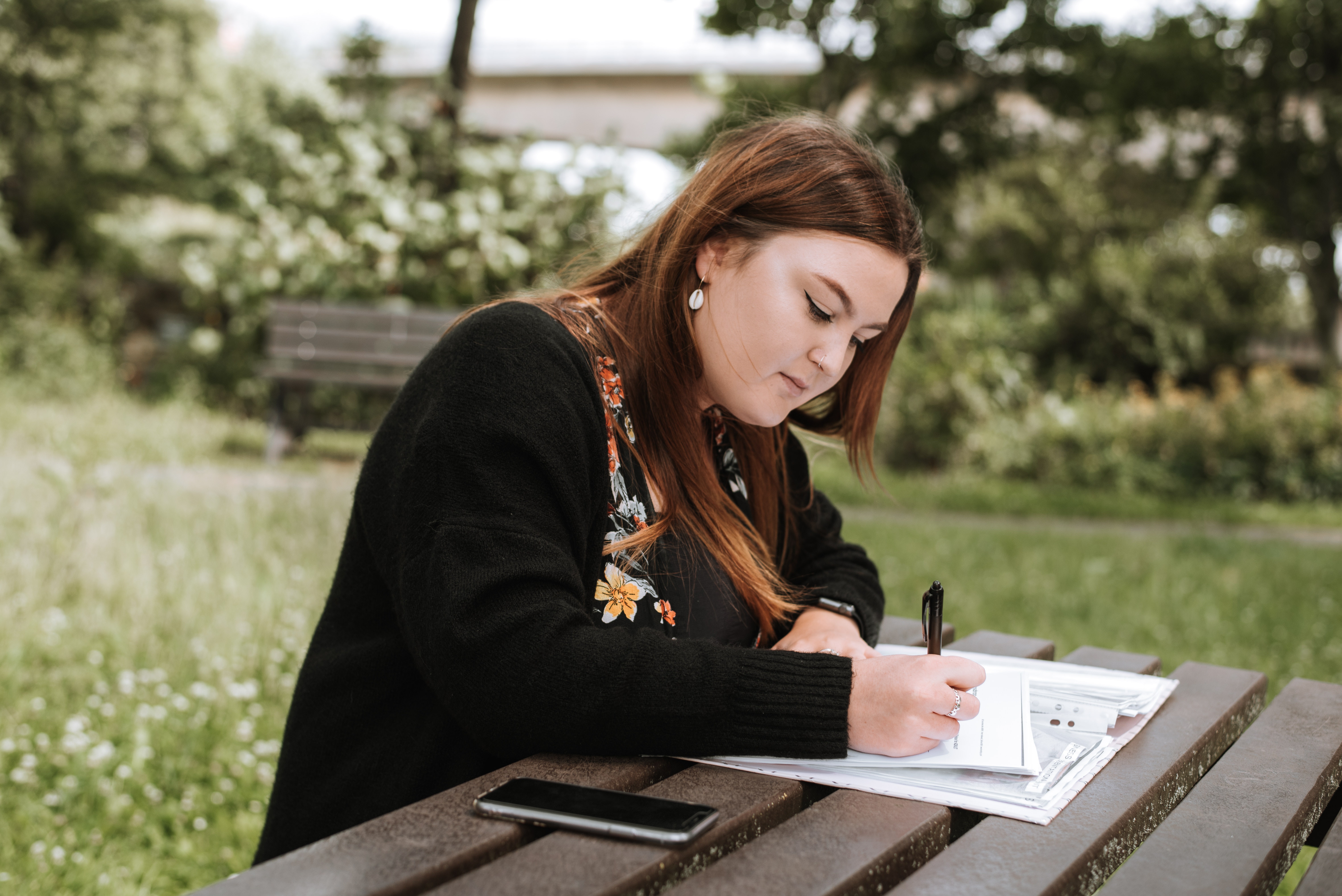 Light skinned woman with long brown hair sits at a picknick bench and writes on papers with a cell phone next to her and a park in the background
