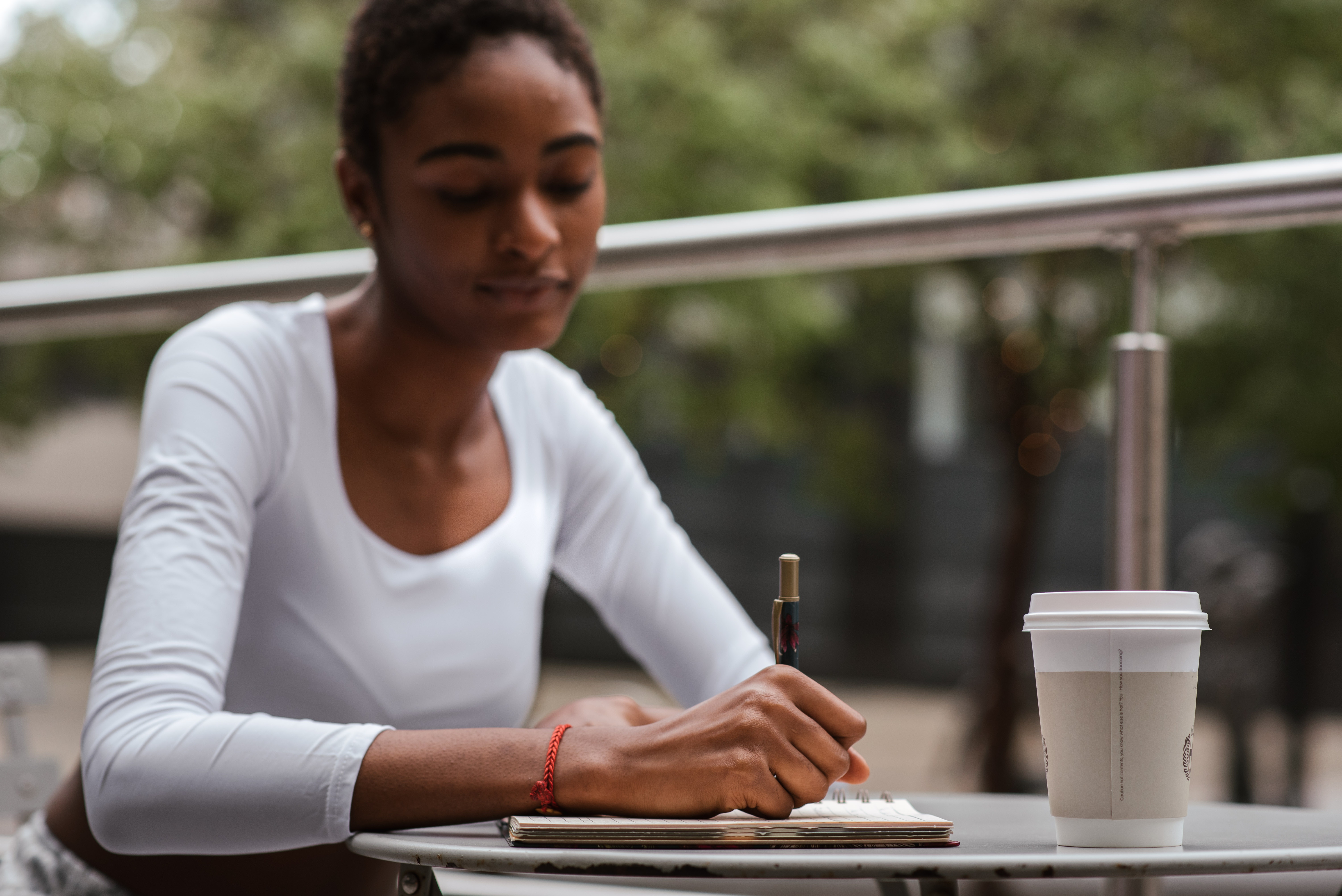 A woman with cark skin and short dark hair sits at a modern outdoor table with a disposable coffee cup writing with a pen