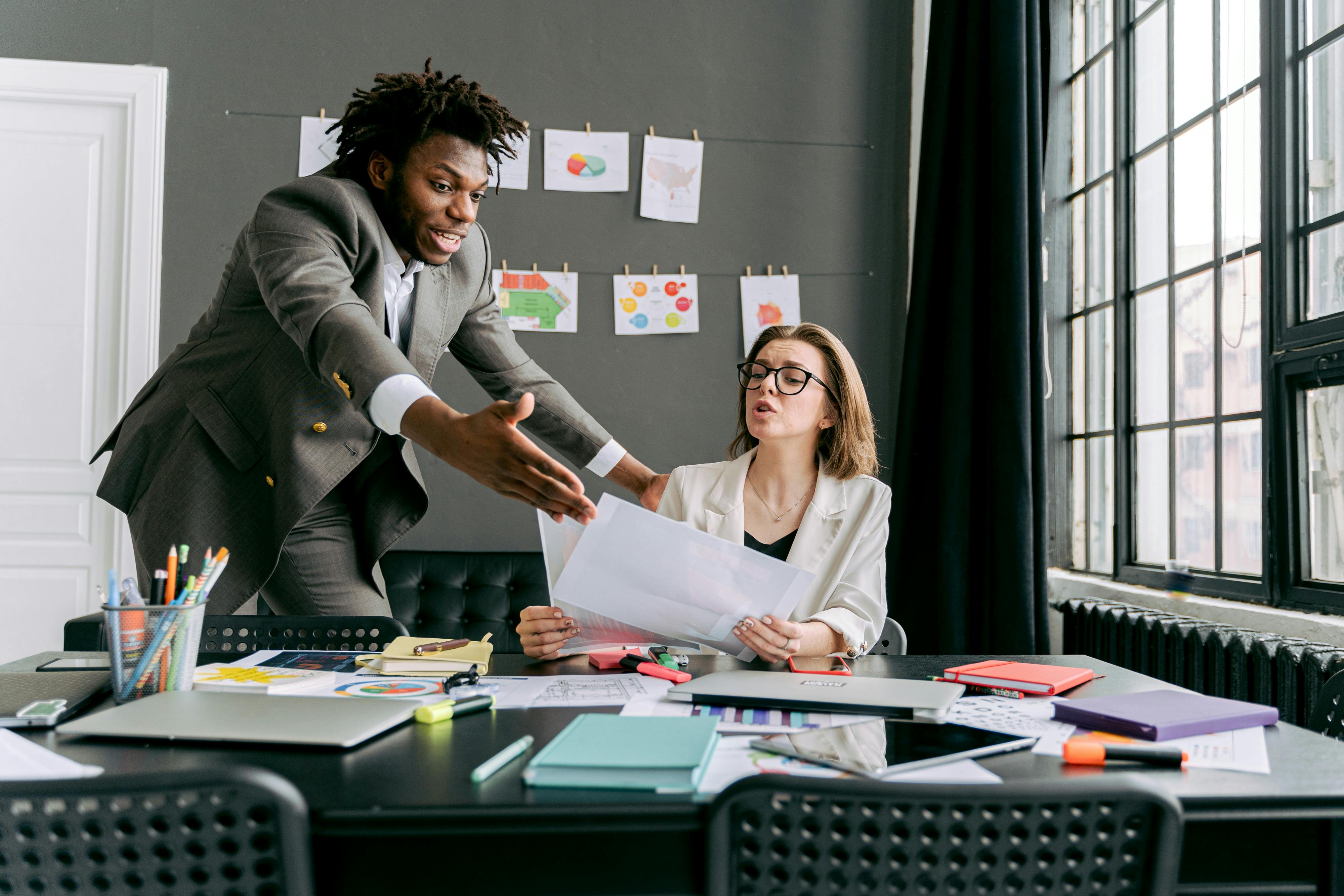 Business man stands gesturing at a paper to business woman sitting at a desk