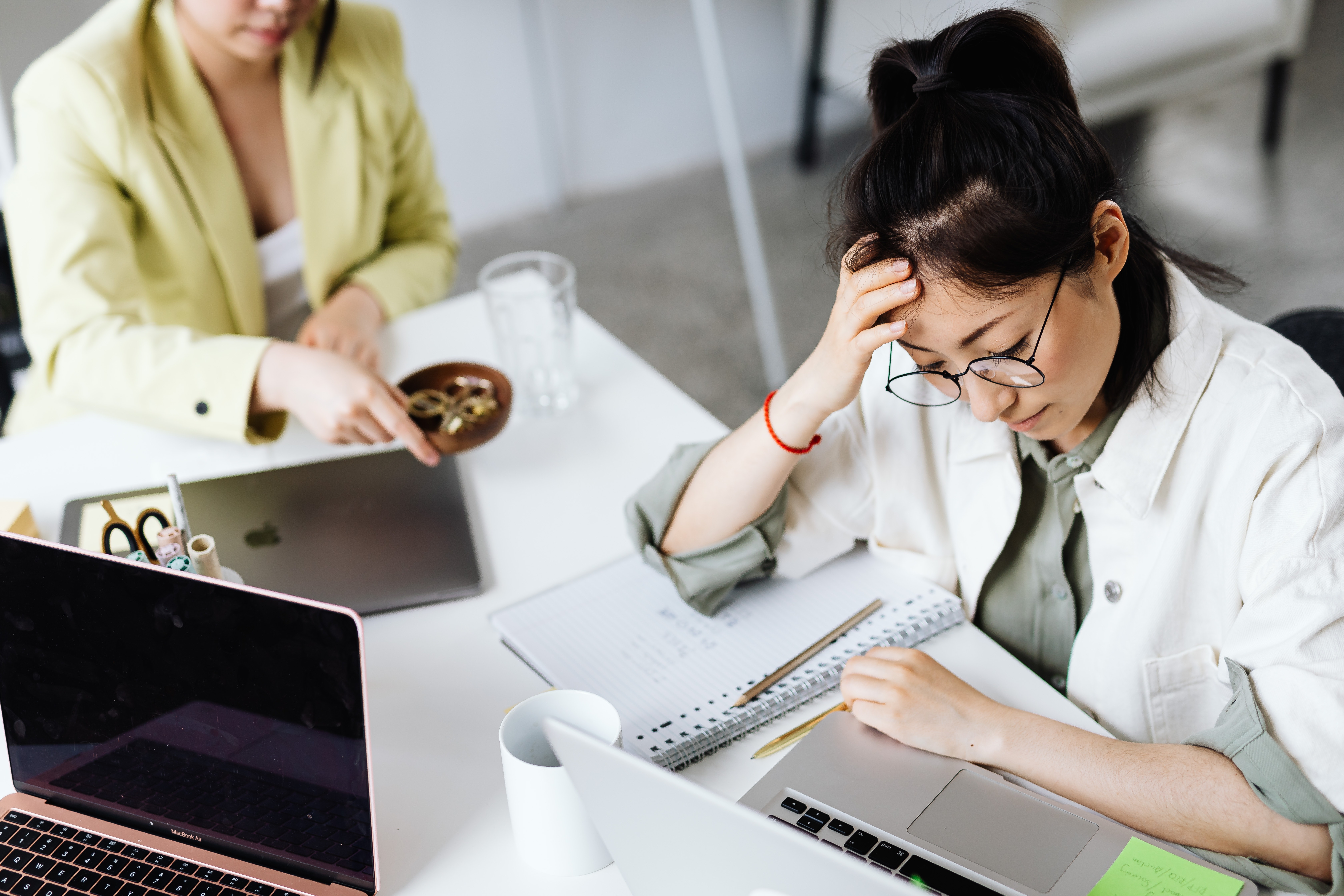 A woman with dark hair and glasses looks exasperated staring at a laptop. There is another person at a different laptop at the table beside her 