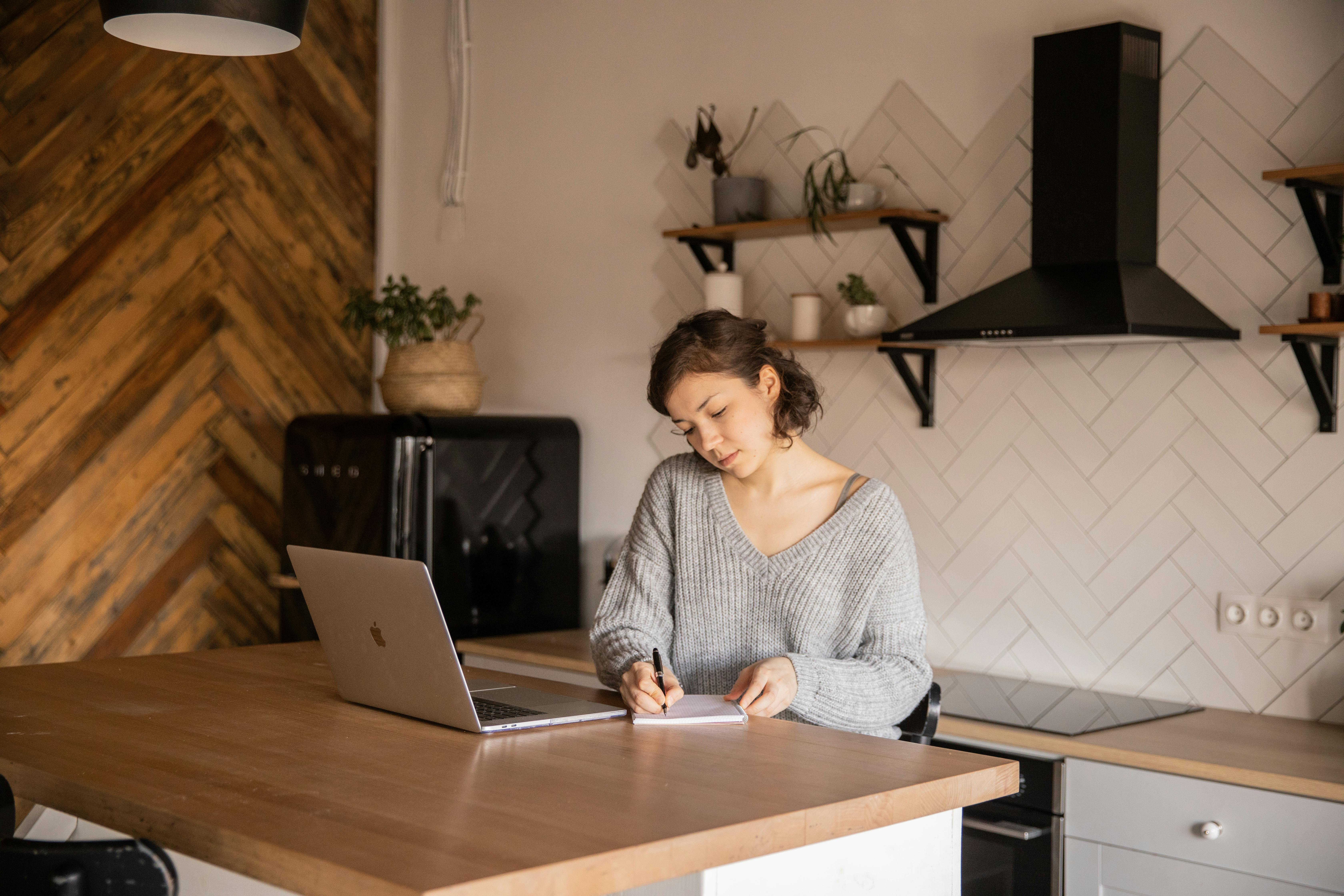 A light skinned woman sits writing on a paper behind a laptop at a kitchen island