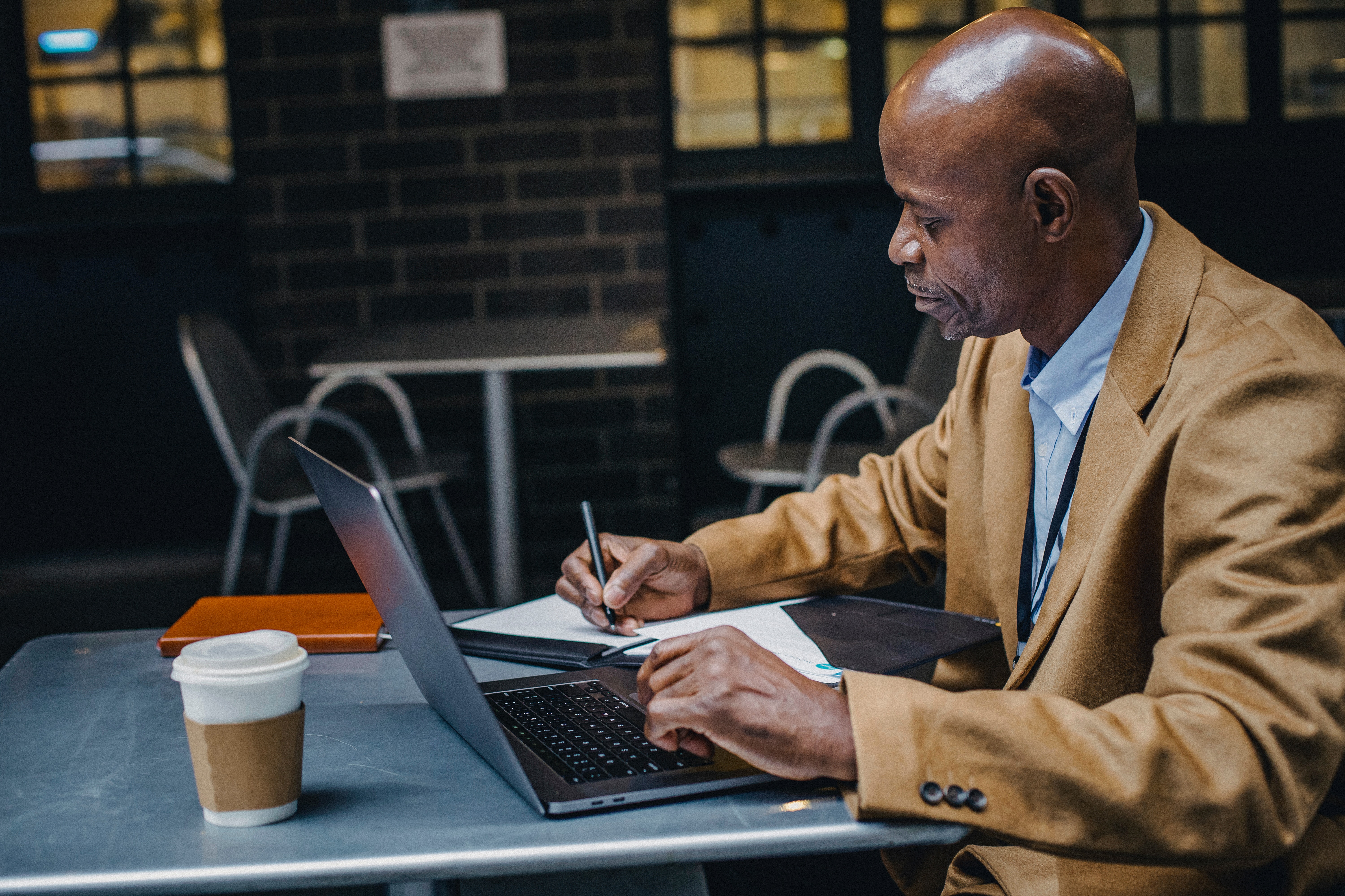 Man with dark skin writes notes with his right hand while sitting at a cafe and working on a laptop. Coffee sits on the table behind the computer.