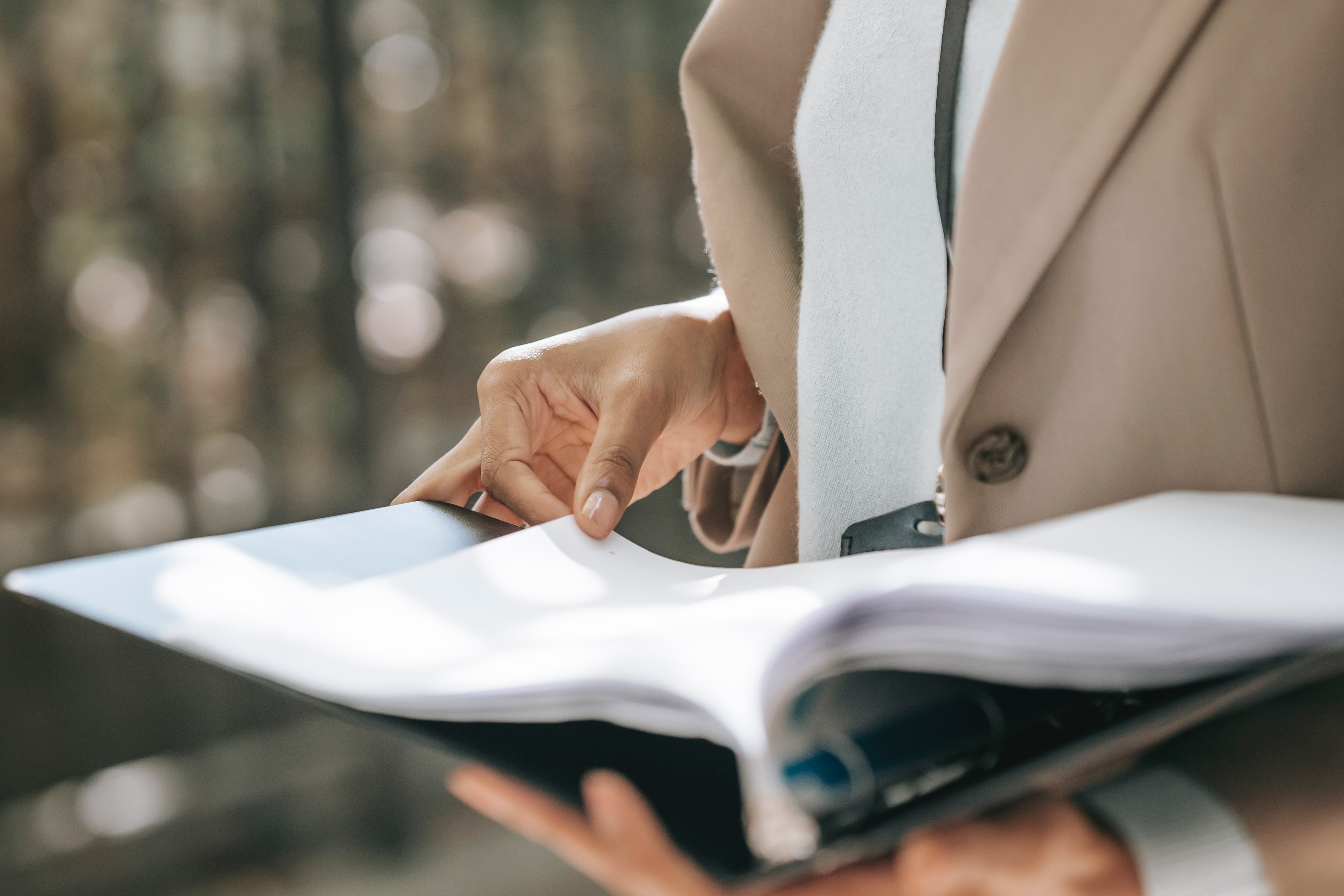 A person with medium skin holds a book open in front of their body in front of a blurry outdoor background