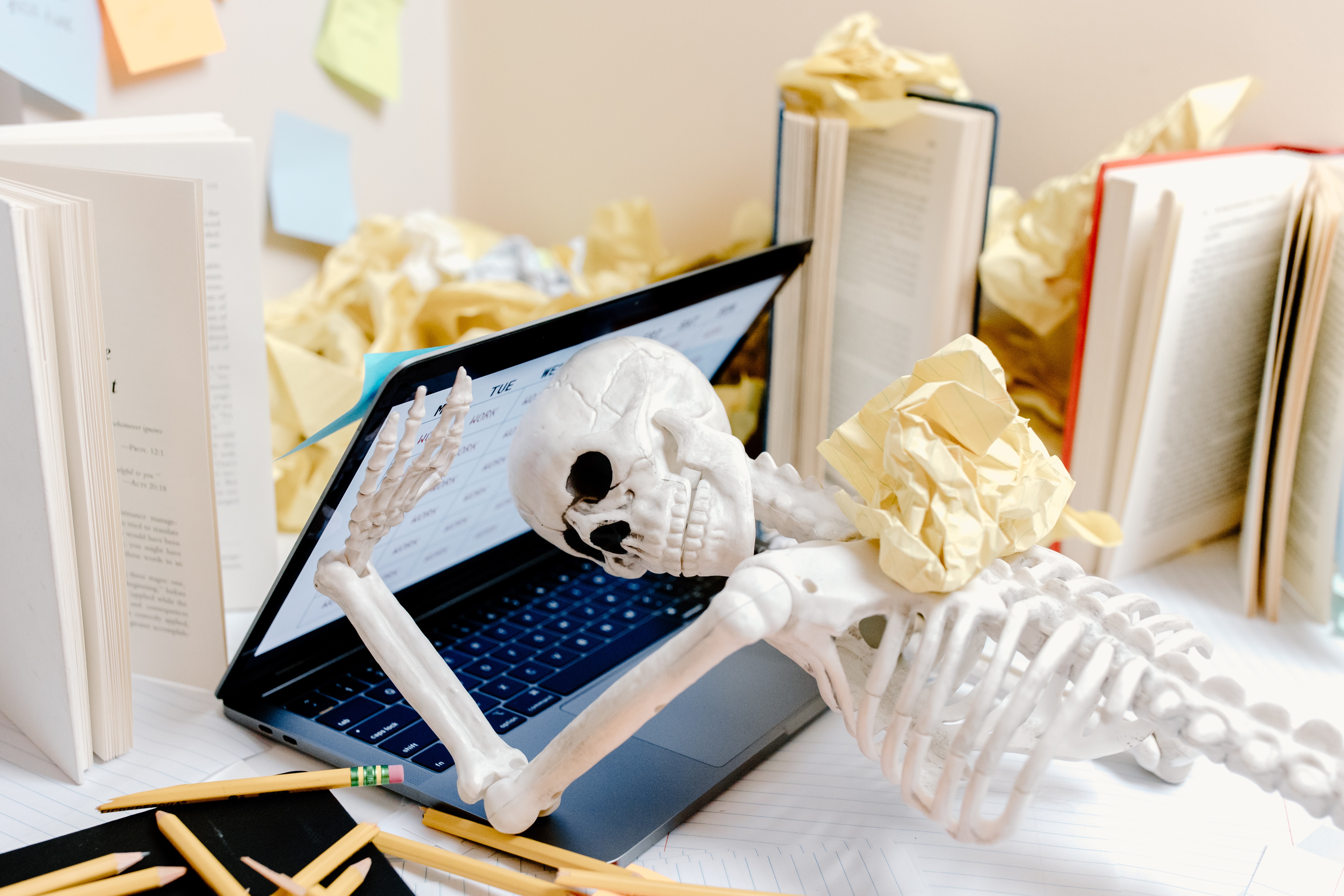 A skeleton has collapsed typing at a laptop with crumpled papers all around it